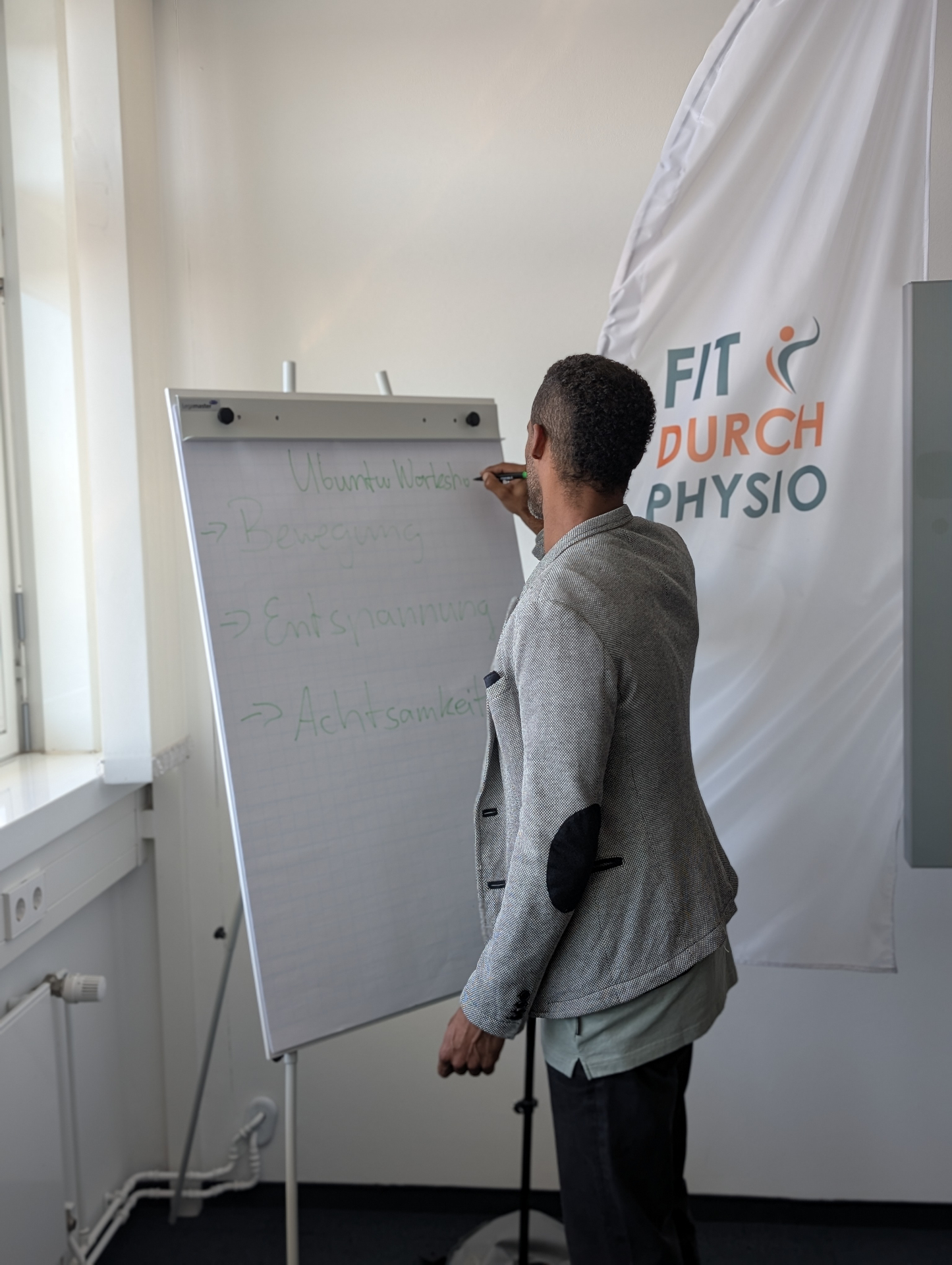 Ubuntu Workshop bei Fit durch Physio: Daniel Günther erklärt die Säulen Bewegung, Entspannung und Achtsamkeit an einem Flipchart vor dem Praxis-Banner.