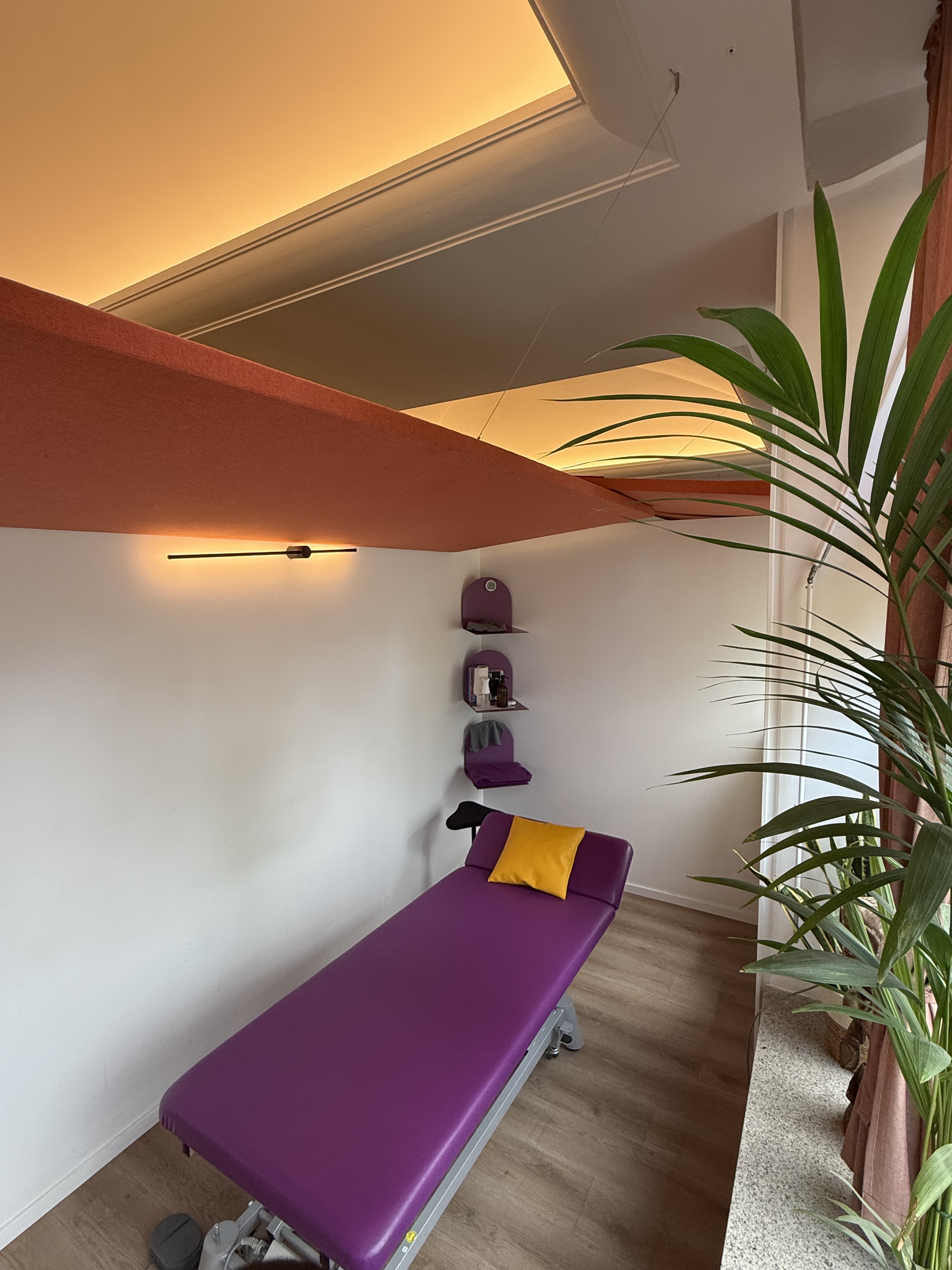 Lila Therapieliege in einem hellen Behandlungsraum von Fit durch Physio mit warmem Deckenlicht, Pflanzen und dekorativen orangefarbenen Deckensegeln.